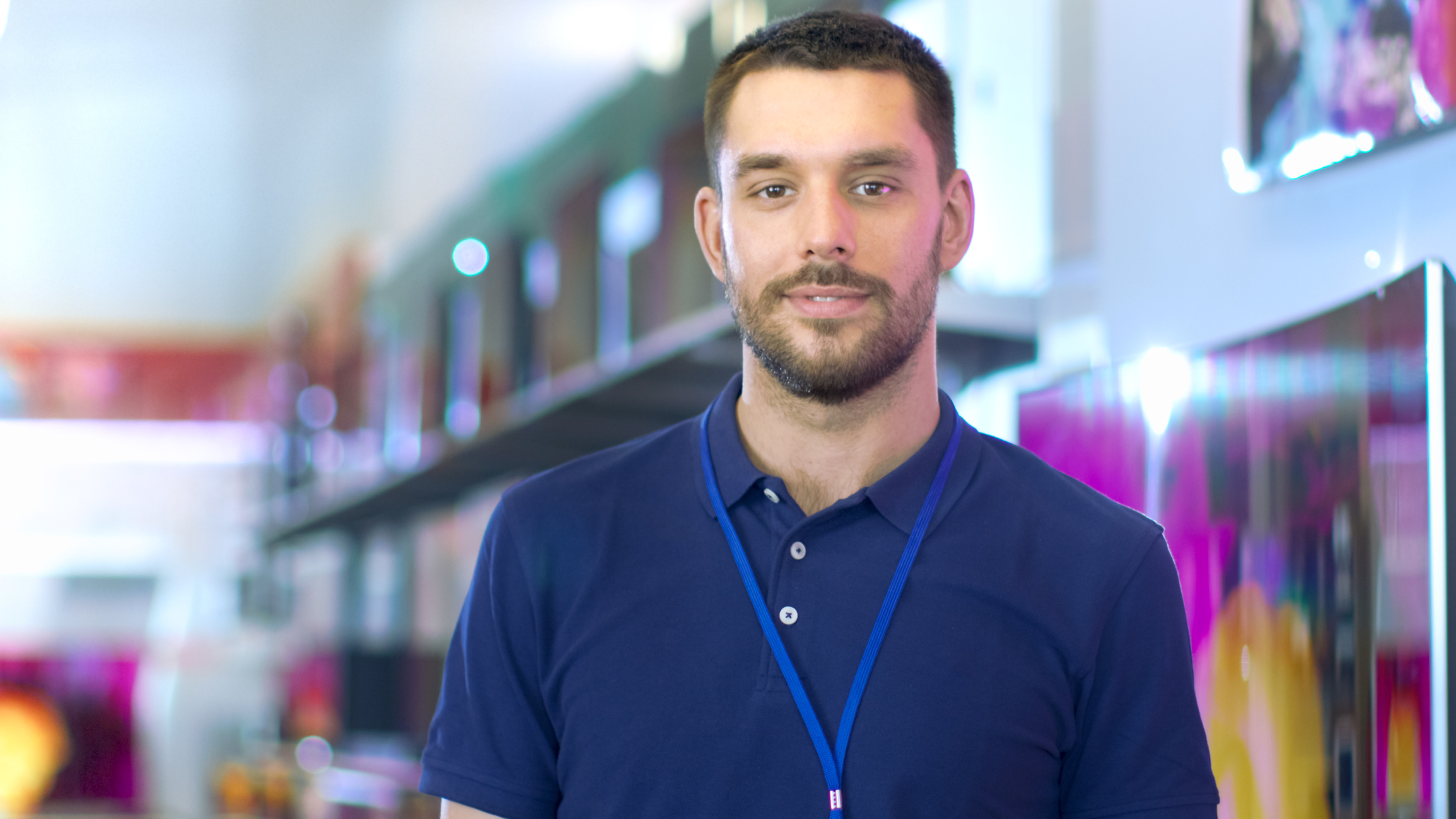 Portrait of a Professional Expert Consultant Smiles and Looks into Camera as Stands in the Bright, Modern Electronics Store Full of Latest Models of TV Sets, Cameras, Tablets and other Devices.The Depth of Field Shot.